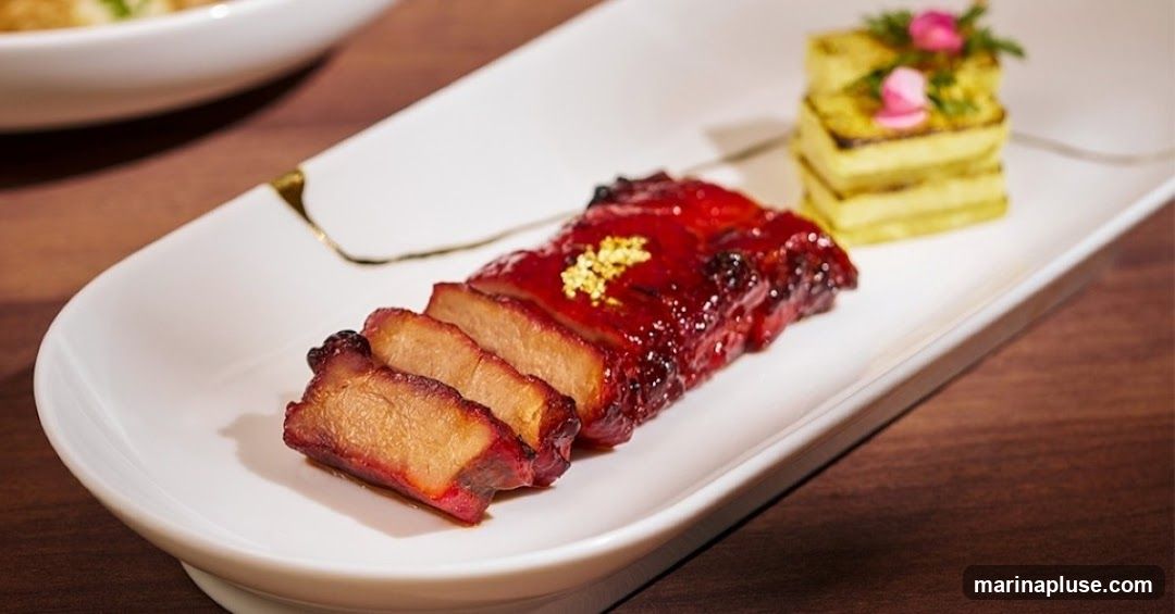 A close-up shot of sliced char siu pork, glazed with a dark red sauce and sprinkled with gold leaf, served on a white platter. To the right, a small stack of layered, grilled yellow food, possibly potato or polenta, is garnished with pink flower petals and green herbs. The platter has a subtle gold trim.