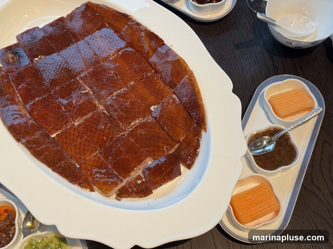 A platter of sliced Peking duck with crispy skin is presented on a white serving dish. Alongside, there are small dishes containing what appears to be fruit slices and a dipping sauce. The meal is set on a dark wooden table.