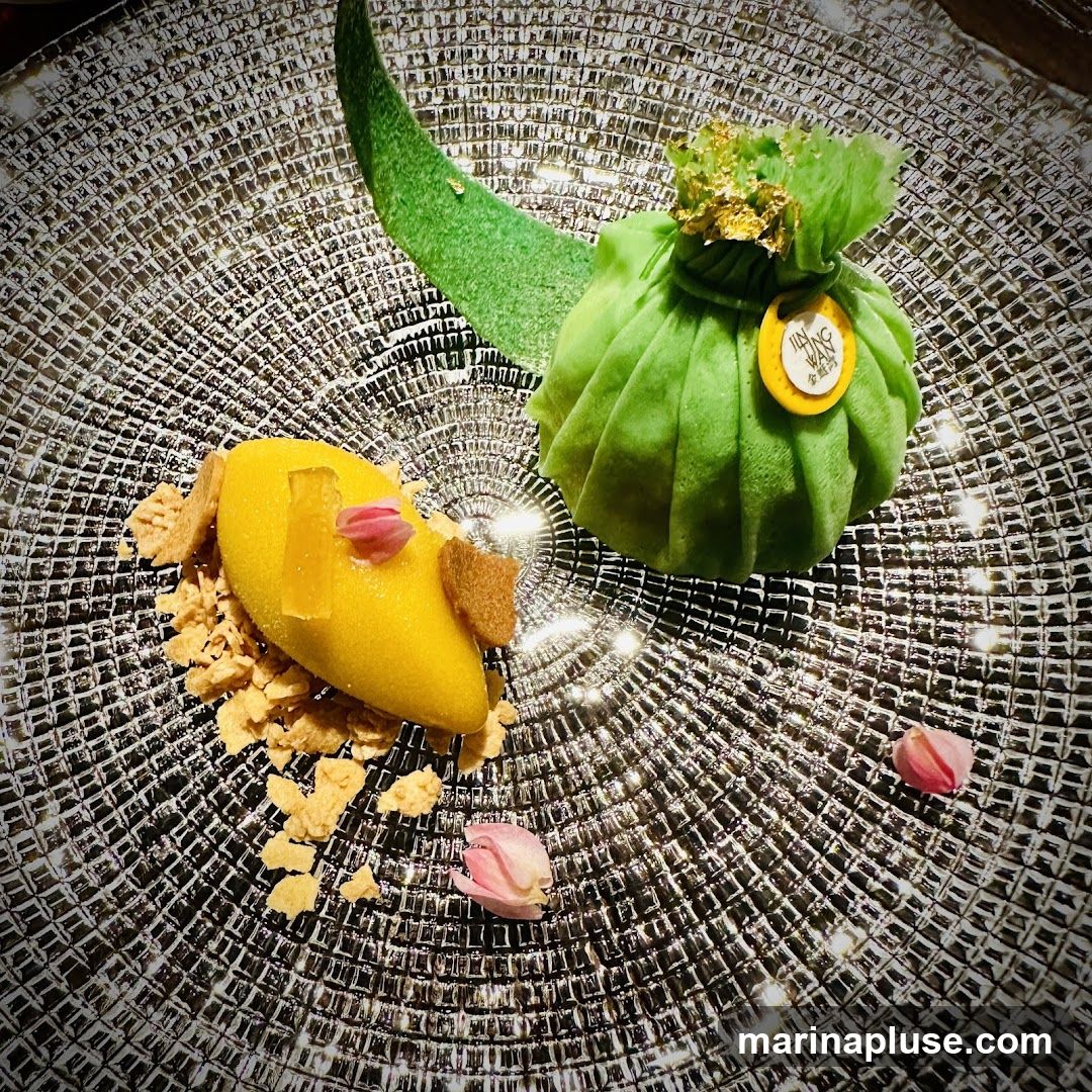 A close-up shot of a dessert served on a textured glass plate. The dessert features a bright yellow mango sorbet, garnished with crumbled cookies, a piece of candied fruit, and delicate pink flower petals. To the right, a green pastry is shaped like a wrapped gift, topped with gold leaf and a small yellow tag with Chinese characters. A green leaf-shaped garnish is placed behind the pastry.