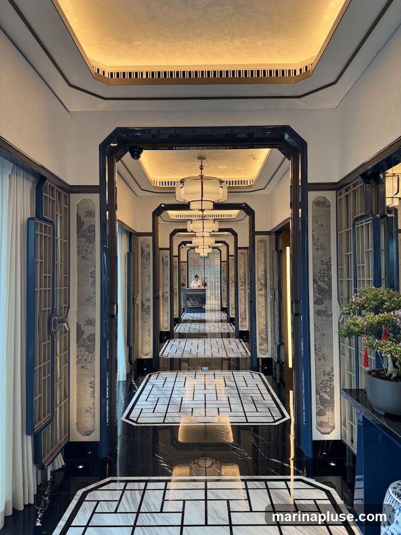 A long, ornate hallway with a repeating pattern of decorative panels and lighting fixtures. The floor is a glossy black marble with white geometric inlays, and the walls feature intricate wallpaper. A bonsai tree sits on a blue cabinet to the right. In the distance, a person can be seen at a reception desk.