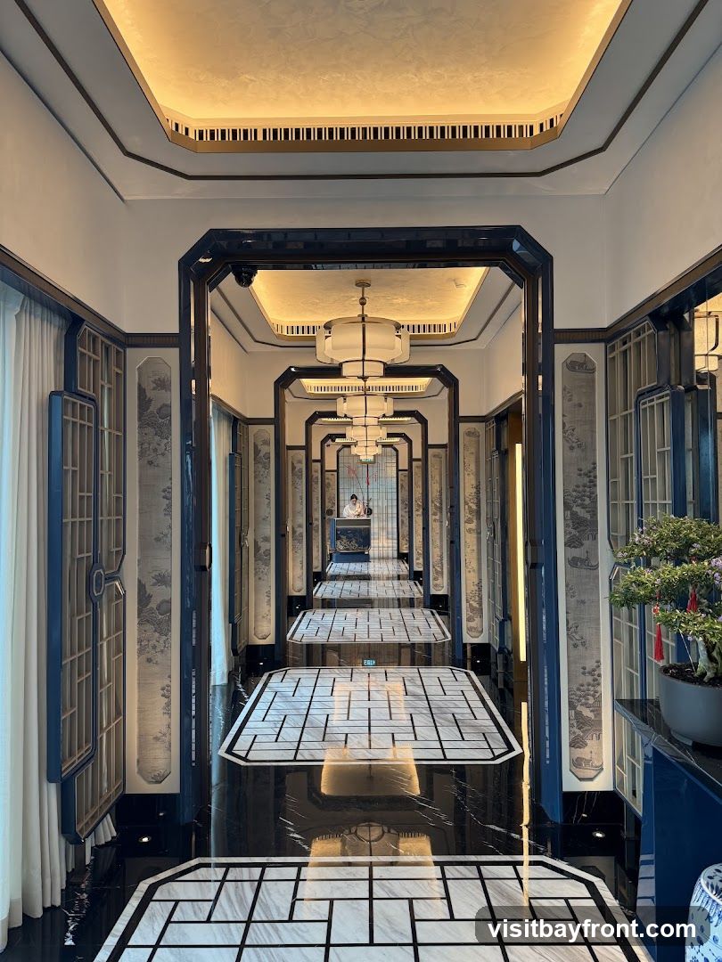 A long, ornate hallway with a repeating pattern of decorative panels and lighting fixtures. The floor is a glossy black marble with white geometric inlays, and the walls feature intricate wallpaper. A bonsai tree sits on a blue cabinet to the right. In the distance, a person can be seen at a reception desk.