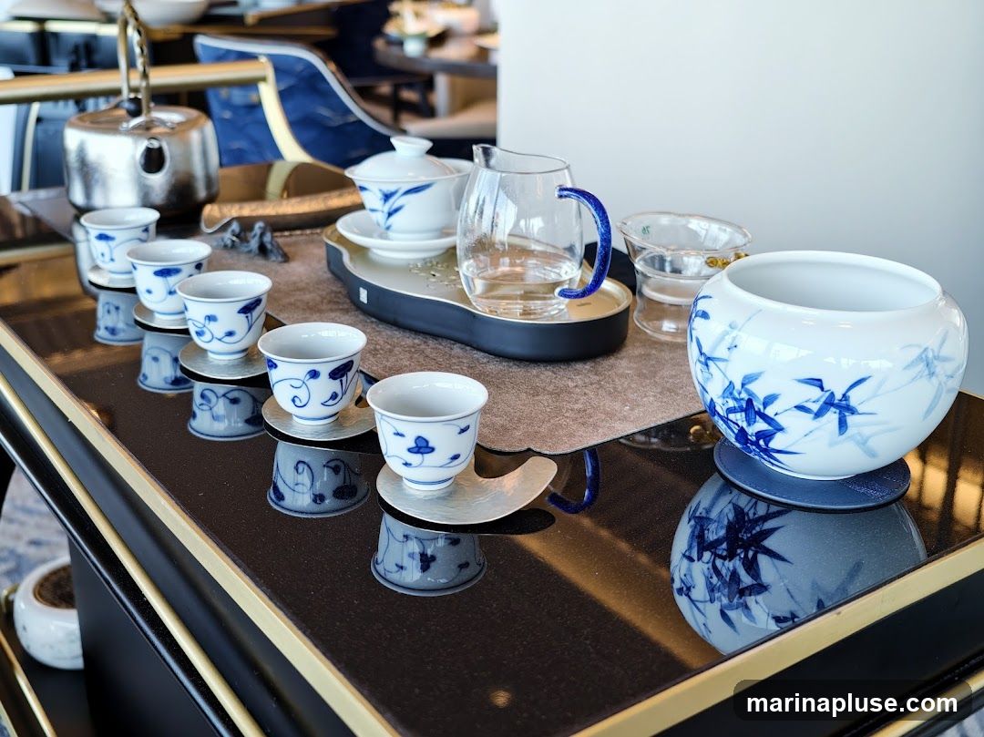 A tea set with multiple small cups and saucers, a glass pitcher with a blue handle, a teapot, and a large decorative bowl, all adorned with blue floral and bamboo patterns. The set is arranged on a dark, reflective surface, likely a table, with a blurred background of a dining area.
