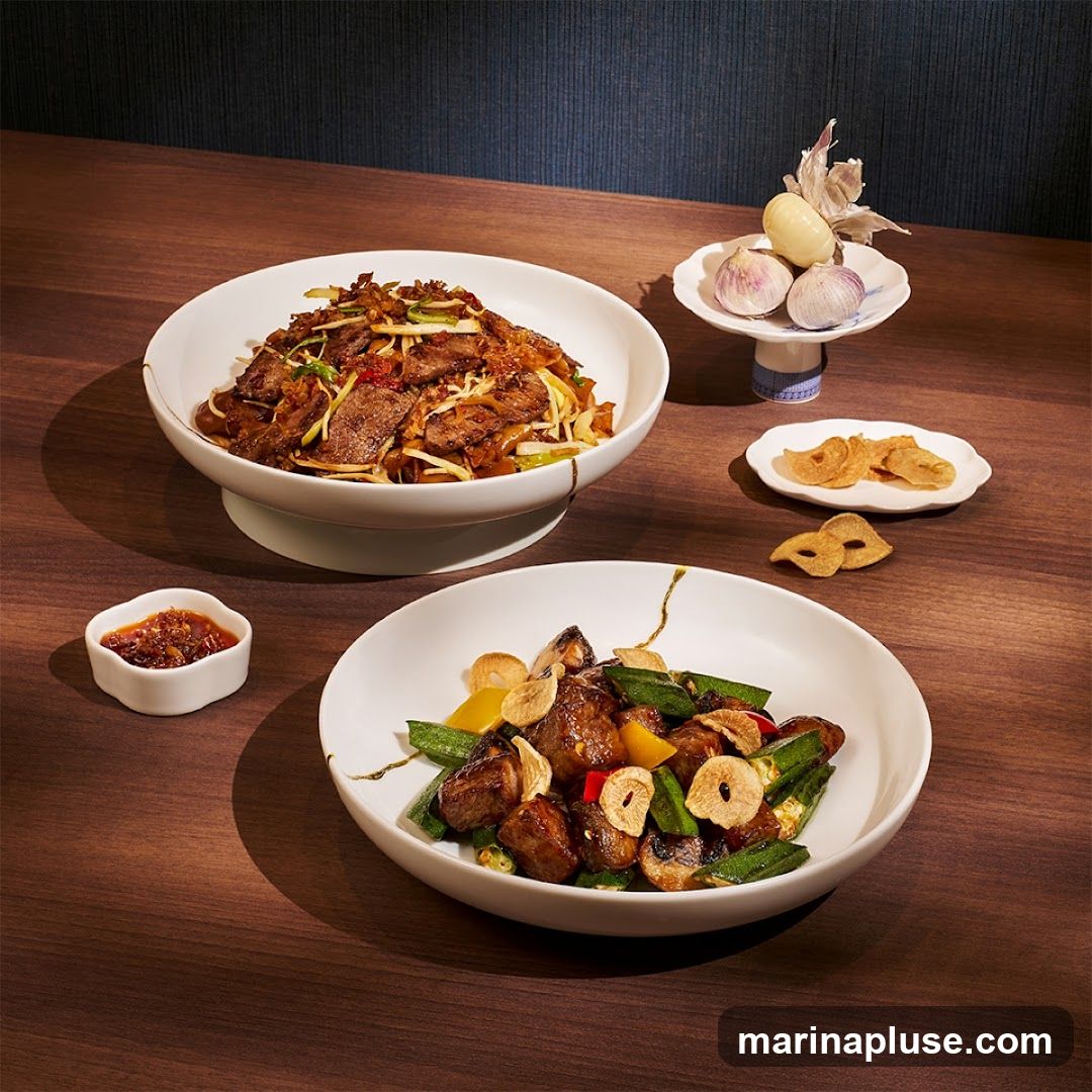 Two bowls of Asian food are presented on a wooden table. The bowl in the foreground contains meat, okra, mushrooms, and garlic slices. The bowl in the background contains noodles with sliced beef and vegetables. A small dish of chili paste and a decorative arrangement of garlic bulbs are also visible.