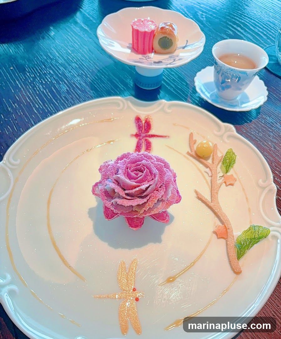 A white plate holds a pink rose-shaped dessert dusted with sugar, two decorative dragonflies, and a small branch with leaves and a yellow orb. A small dish with two pink and green confections sits on a stand in the background, along with a cup of tea.