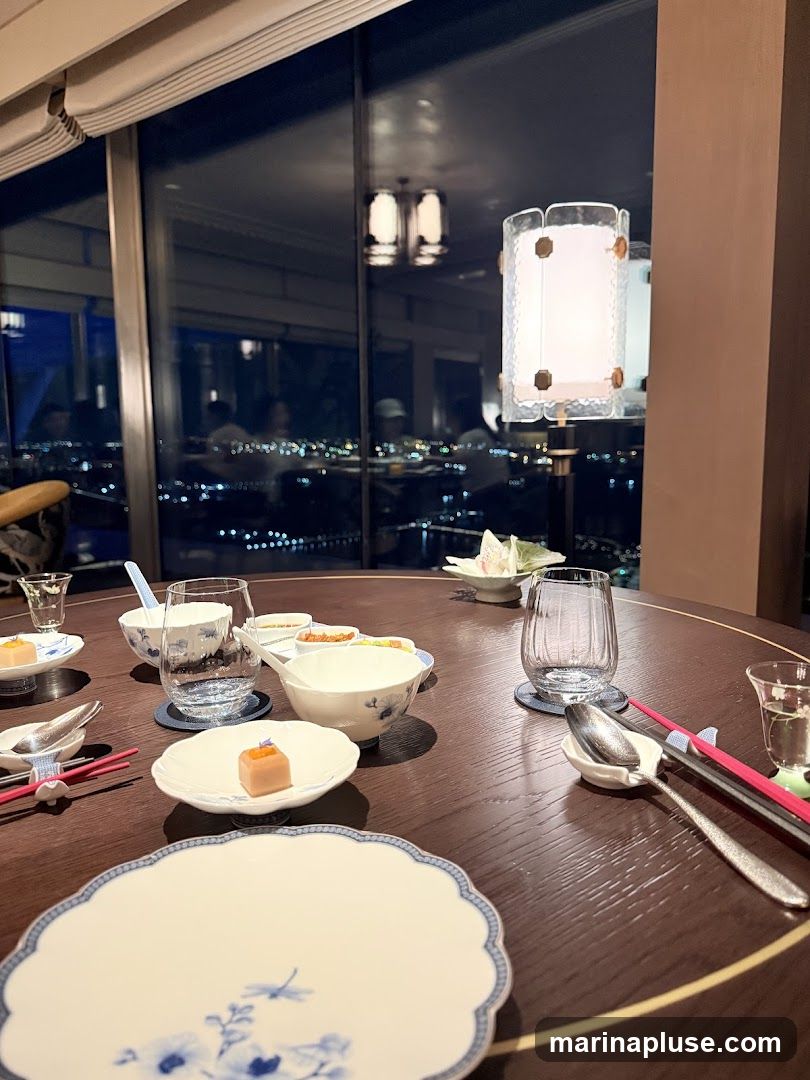 A round, dark wood dining table is set for a meal, overlooking a city at night through a large window. The table features white plates with blue floral designs, small bowls containing appetizers, chopsticks, spoons, and glasses. A decorative lamp hangs above, casting a warm glow. The city lights twinkle in the background, creating a sophisticated ambiance.