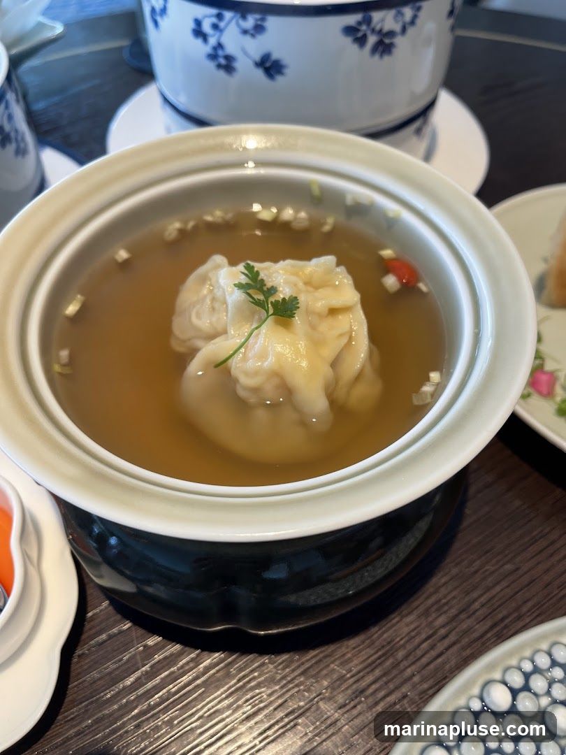 A close-up shot of a bowl of dumplings in a clear broth, garnished with chopped green onions and a sprig of parsley. The bowl is light-colored with a darker base and sits on a wooden table. In the background, a blue and white patterned teapot and lid are visible.