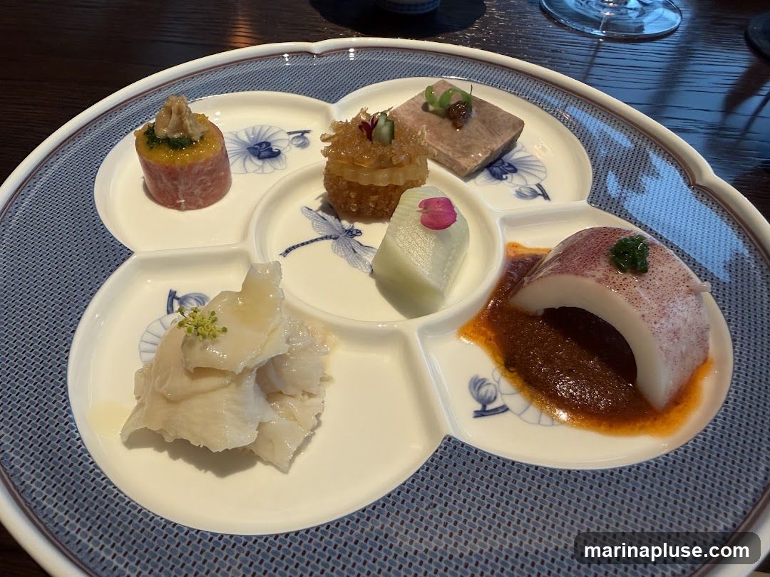 A close-up shot of a multi-compartment plate holding various small appetizers. The plate has a blue and white pattern with dragonflies and floral motifs. The appetizers include a cylindrical piece of meat topped with greens, a small crispy ball, a slice of meat, a piece of white vegetable with a pink flower petal, a portion of white seafood with a light sauce and a garnish, and a piece of squid with a dark sauce.