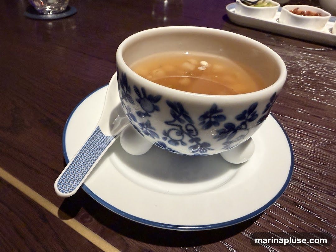 A close-up shot of a bowl of soup with a spoon on a saucer. The bowl is white with blue floral patterns. In the background, there are small dishes containing appetizers. The setting appears to be a restaurant table.
