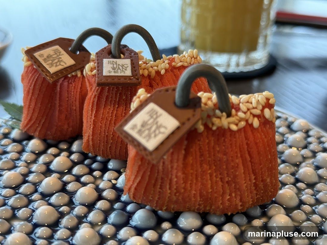 Three orange, pleated appetizers shaped like handbags are presented on a decorative plate with a pattern of blue and white raised dots. Each appetizer has a dark brown handle and a small, square tag attached, with some tags displaying Chinese characters and the text "JIN WANG". The top edges of the appetizers are sprinkled with sesame seeds. A glass of amber liquid is visible in the blurred background.