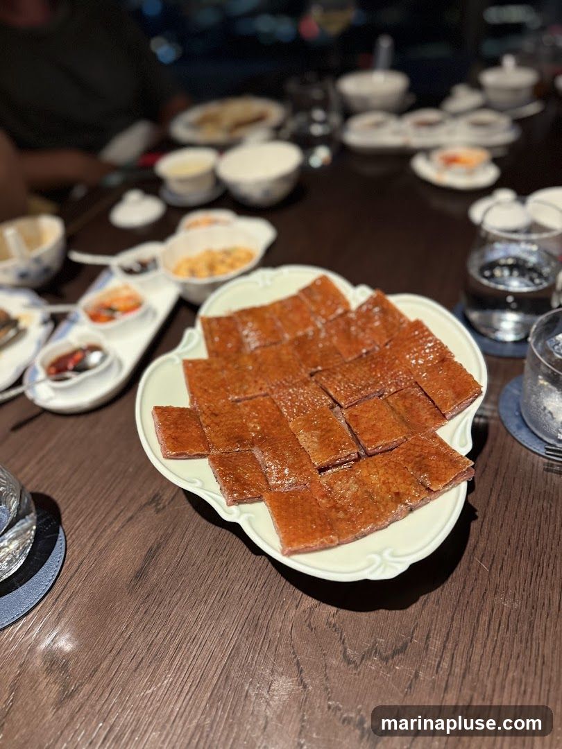 A platter of sliced Peking duck is presented on a table, with various small dishes and glasses of water in the background. The duck skin is crispy and glazed, cut into rectangular pieces.