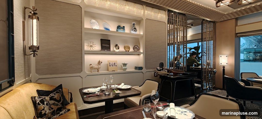 An elegant dining room with a plush yellow sofa, dark wood tables set for dining, and decorative shelves displaying vases and art pieces. A person is visible in the background, appearing to work behind a counter. The ambiance is sophisticated and dimly lit, with modern lighting fixtures and large windows offering a view of the city.