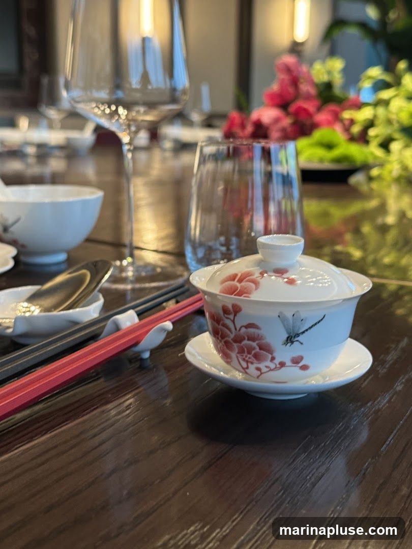 A close-up shot of a table setting with a lidded tea cup adorned with floral and dragonfly motifs, chopsticks resting on a holder, and a spoon in a small dish. In the background, wine glasses, water glasses, and floral arrangements are visible, suggesting a dining or tea ceremony setting.