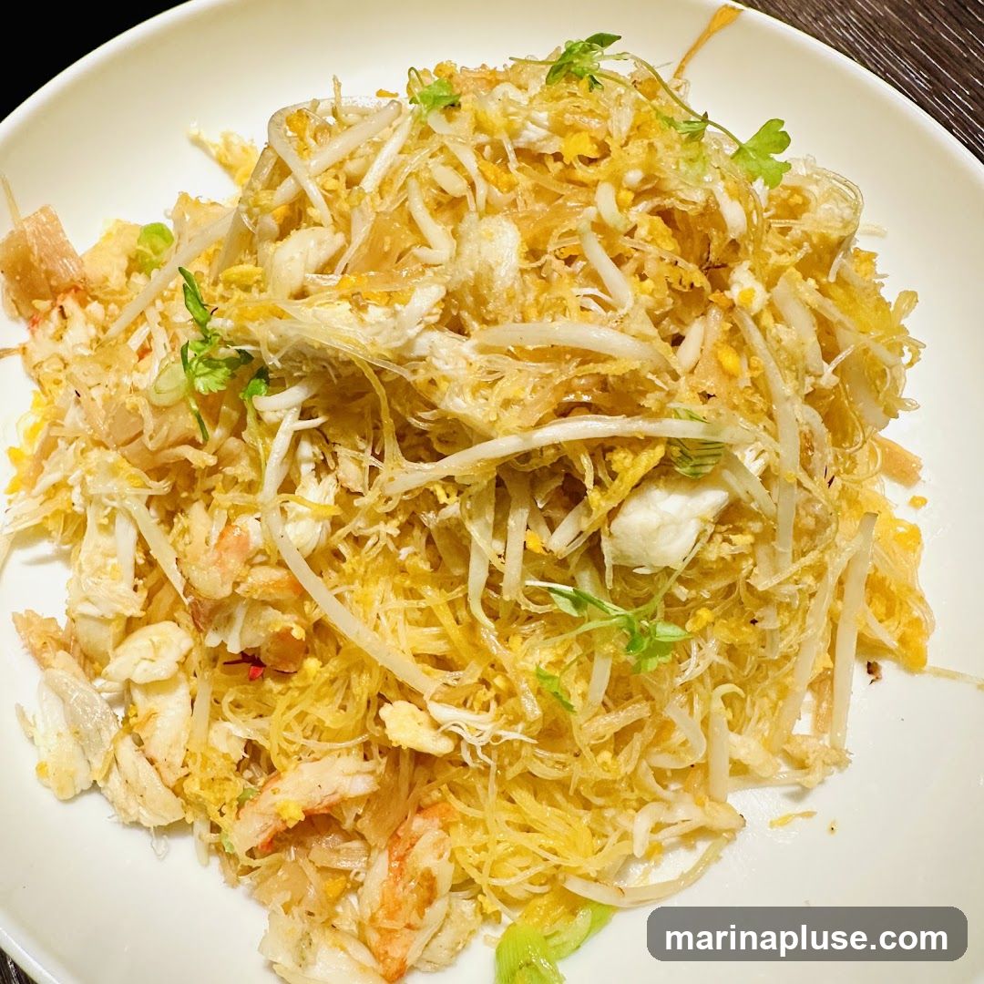 A close-up shot of a white plate filled with a generous portion of stir-fried noodles. The noodles are thin and golden, mixed with shredded crab meat, pieces of shrimp, bean sprouts, and scrambled egg. Garnished with fresh green herbs, the dish appears savory and flavorful.