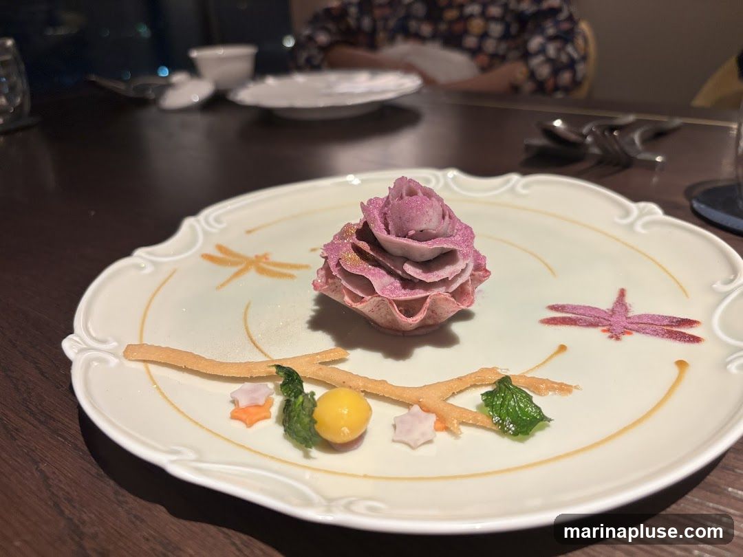 A delicate dessert is presented on a decorative white plate. The dessert is shaped like a pink rose with a swirl pattern, dusted with edible glitter. It sits on a small, edible tart shell. The plate is adorned with painted dragonflies and a twig-like garnish. Small decorative elements, including star-shaped candies and a green leaf, are arranged around the twig. The setting appears to be a restaurant with a dark wooden table and blurred background elements suggesting other diners and tableware.