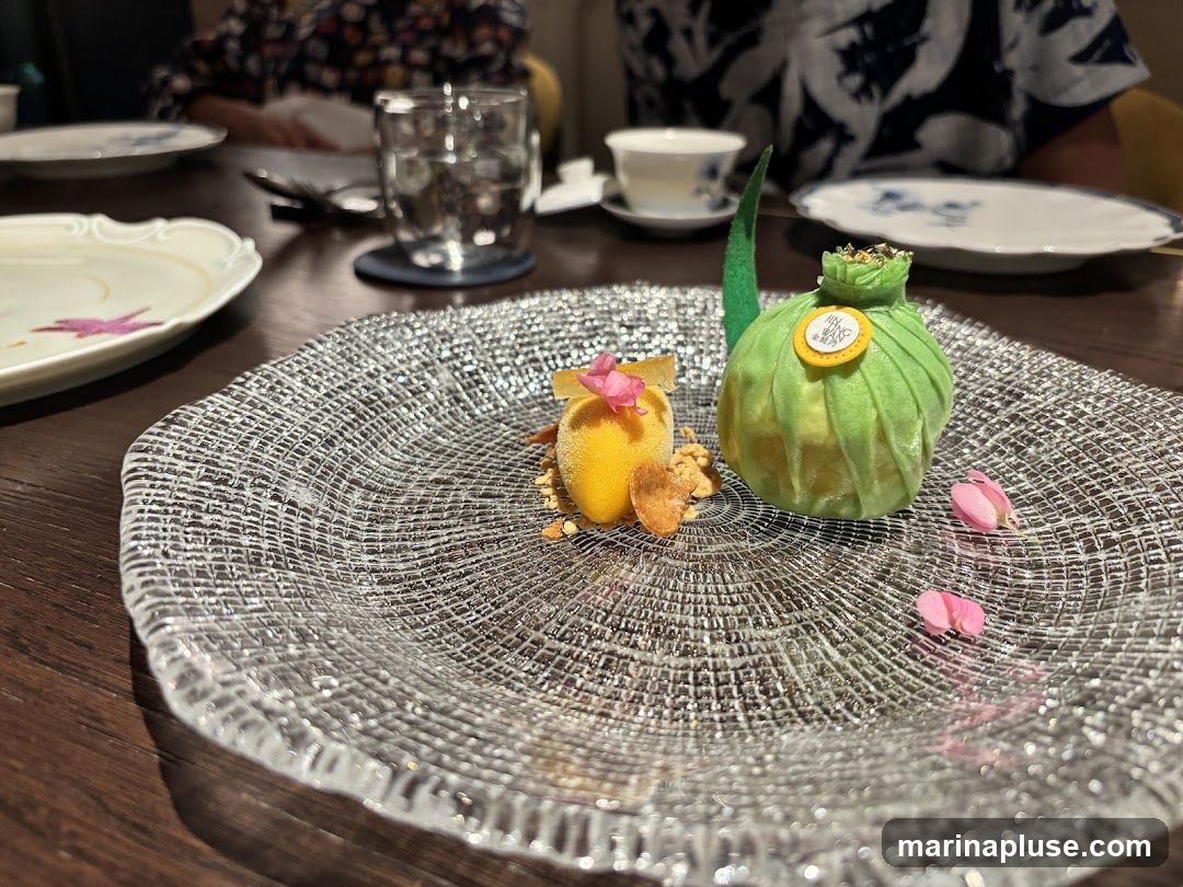 A close-up shot of a dessert presented on a textured glass plate. The dessert features a green, pouch-like pastry with a yellow circular emblem on top, and a small yellow confection with a pink flower garnish. Edible gold flakes are sprinkled on top of the green pastry. The plate is set on a dark wooden table, with other plates and a glass of water visible in the background.