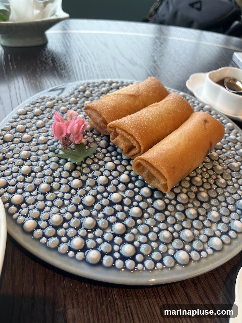 Three golden-brown spring rolls are arranged on a textured blue and white plate. A small pink flower with a green leaf is placed to the left of the spring rolls. A small white dish with a dark sauce and a spoon is visible in the upper right corner. The plate sits on a dark wooden table.