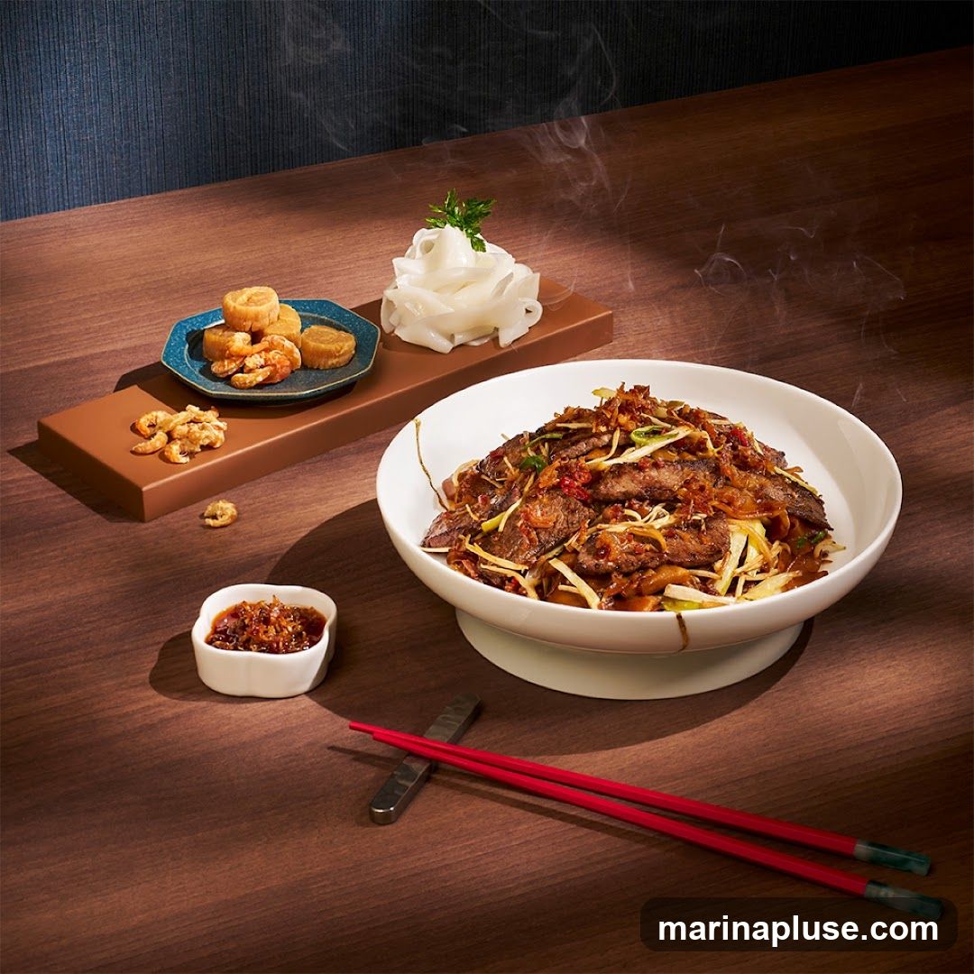 A steaming bowl of beef chow fun, a popular Chinese noodle dish, is presented on a white pedestal bowl. The noodles are wide and flat, mixed with thinly sliced beef, scallions, and crispy fried shallots. To the side, a small dish holds a spicy chili sauce, and a separate plate features dried shrimp and what appear to be small fried cakes. A stack of fresh rice noodles, garnished with parsley, is also visible. Red chopsticks rest on a metal chopstick holder, completing the appetizing presentation.