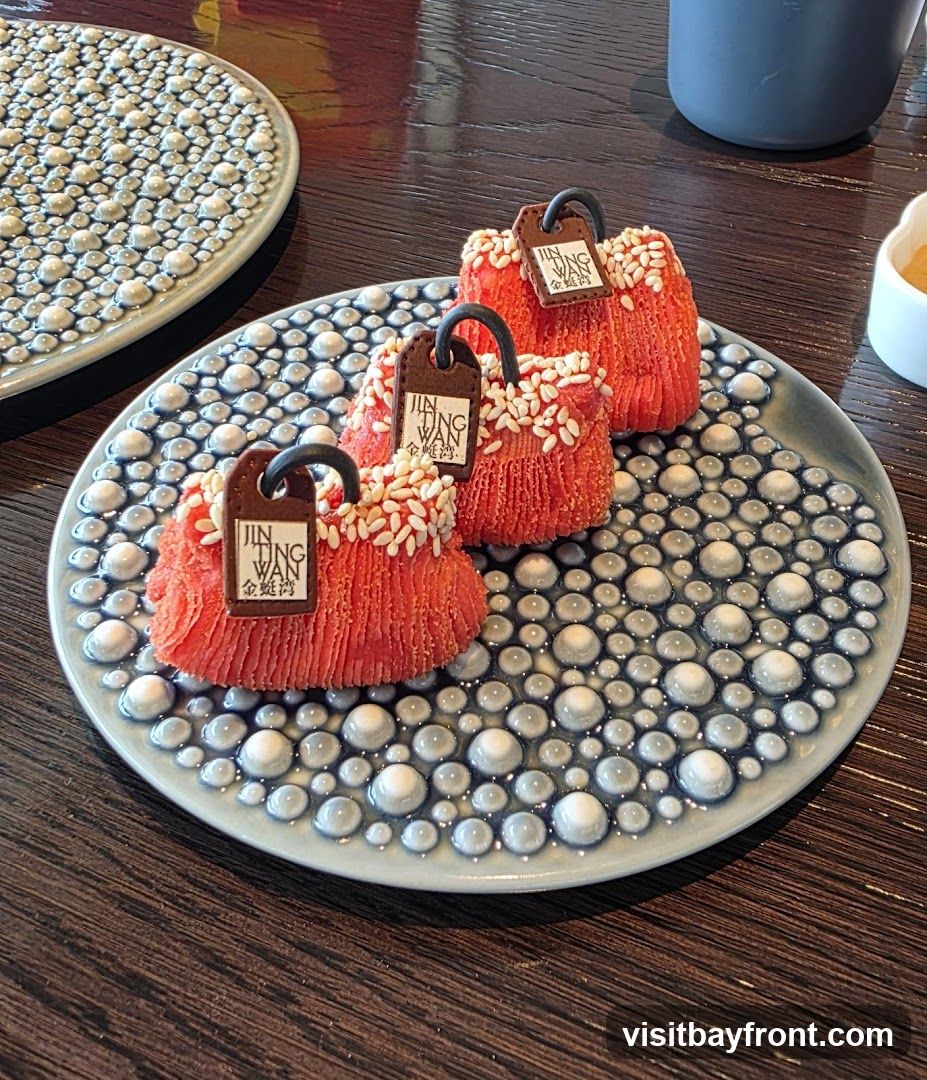 Three red, textured desserts resembling handbags are arranged on a decorative plate. Each dessert has a dark brown tag with the text "JIN TING WAN" and Chinese characters. The desserts are sprinkled with white sesame seeds. The plate has a raised, bubbly pattern in blue and white. Another plate with a similar pattern is visible in the background.