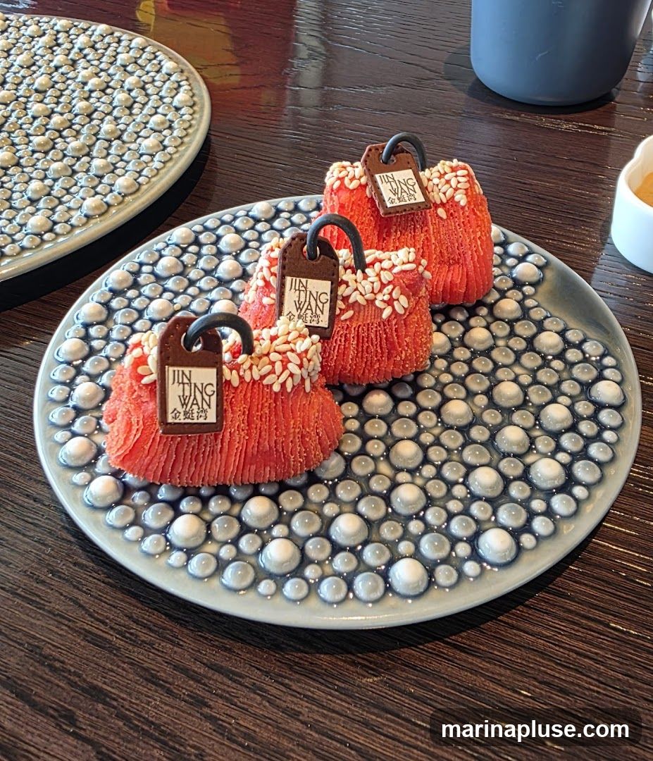 Three red, textured desserts resembling handbags are arranged on a decorative plate. Each dessert has a dark brown tag with the text "JIN TING WAN" and Chinese characters. The desserts are sprinkled with white sesame seeds. The plate has a raised, bubbly pattern in blue and white. Another plate with a similar pattern is visible in the background.