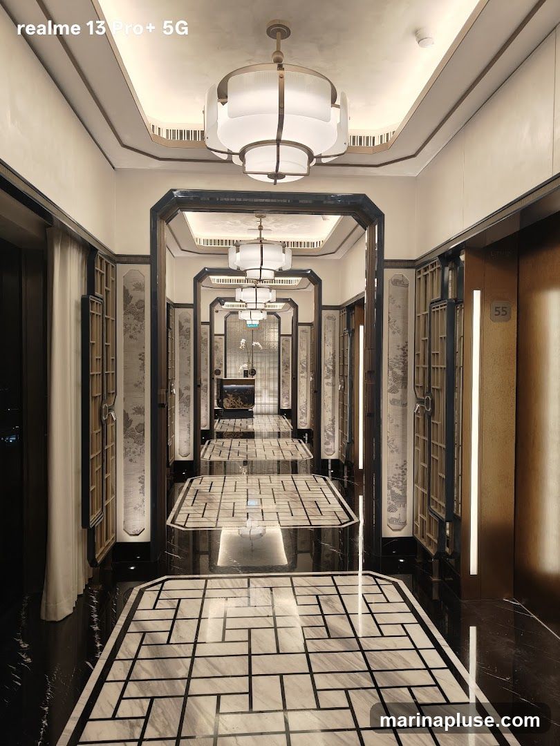 A long, ornate hallway with a repeating pattern of decorative ceiling lights and framed wall panels. The floor is a polished black marble with inlaid geometric patterns. Elevators are visible on the right side.