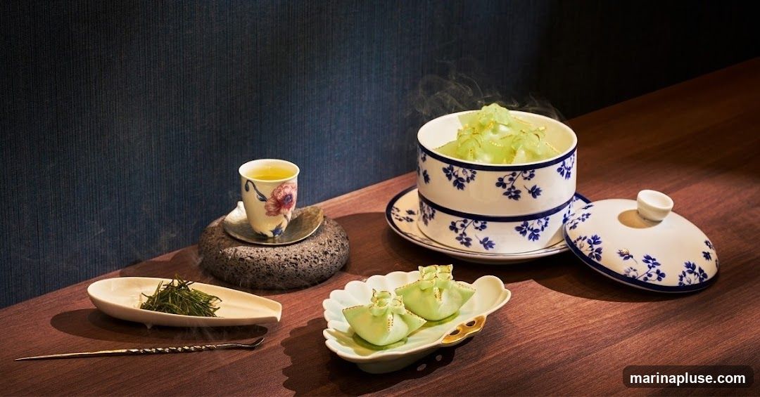 A delicate presentation of a steamed green dessert, possibly mochi or dumplings, served in a lidded porcelain bowl with blue floral accents. Beside it, a cup of green tea sits on a dark, textured coaster. A small dish holds dried green tea leaves, and a long, slender utensil rests on the wooden surface. The background is a dark, textured wall, and the scene is lit to highlight the food and drink.
