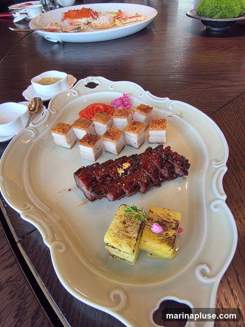 A platter of roasted pork belly with crispy skin, slices of char siu (barbecued pork), and two pieces of layered potato or taro, garnished with pink flowers and herbs. In the background, a plate with king crab legs and a small dish with green moss are visible.