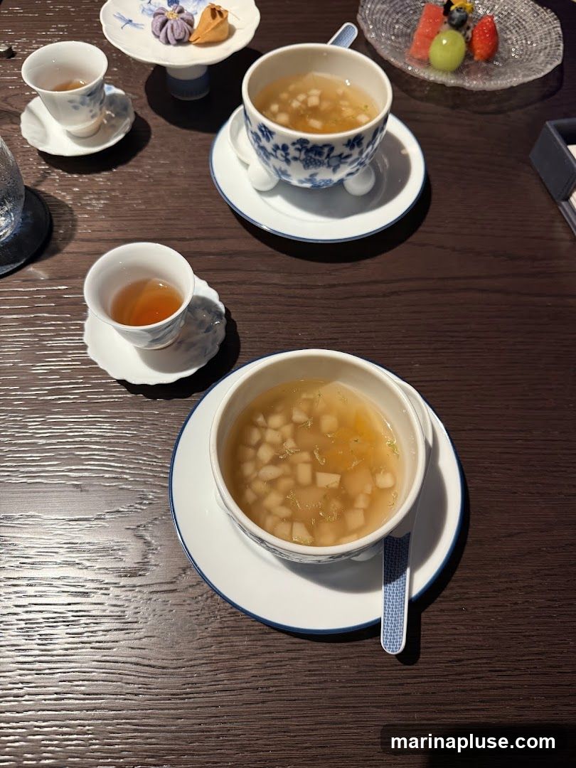 A close-up overhead view of a table setting with two cups of tea, two bowls of soup, a small plate with a pastry and a decorative purple confection, and a small plate of fruit. The table is made of dark wood. The cups and bowls are white with blue floral patterns. One bowl of soup is in the foreground, and another is in the background. One cup of tea is in the foreground, and another is in the background. The fruit plate contains slices of watermelon, a green grape, and a strawberry.