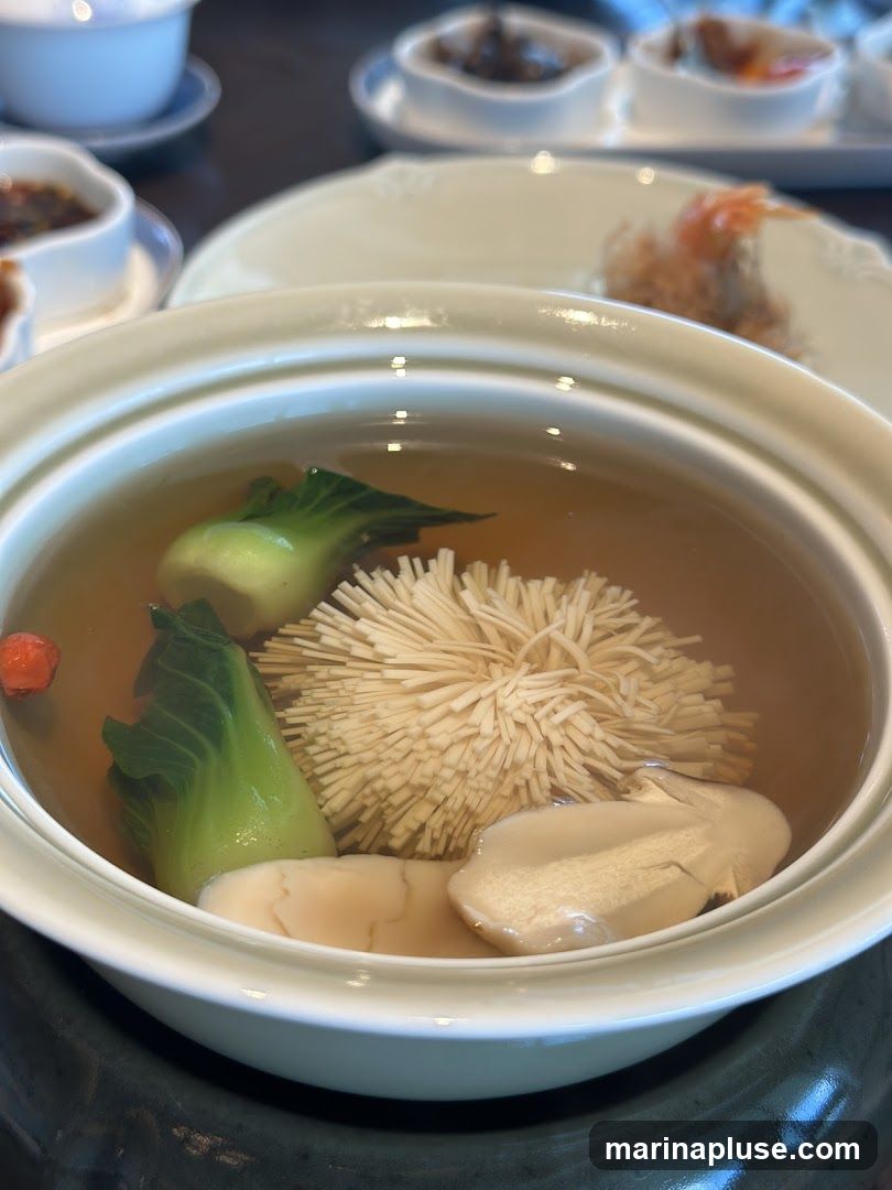 A close-up shot of a bowl of clear broth soup. Inside the soup are pieces of bok choy, enoki mushrooms, and what appear to be slices of abalone or similar seafood. In the background, out of focus, are small dishes containing other food items and a white bowl.