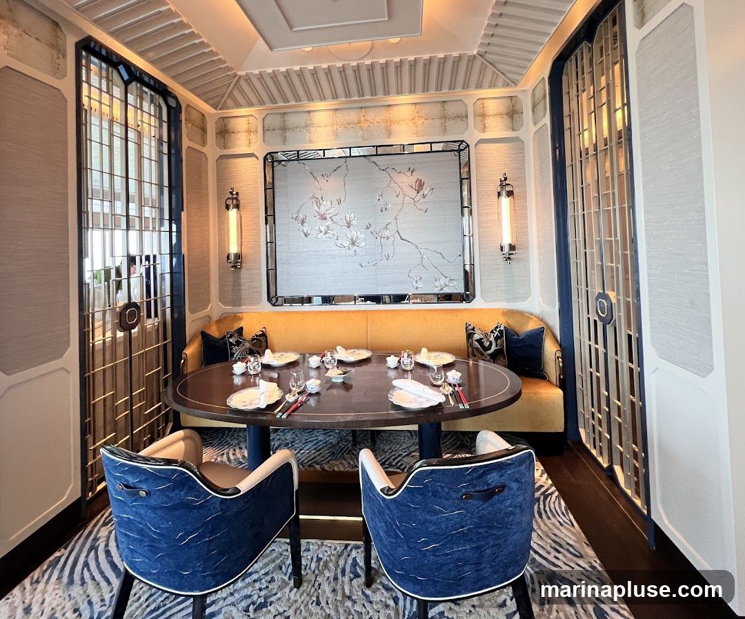 A beautifully set dining table in a luxurious room with blue and gold accents. The table is round and dark wood, set with white plates, cutlery, and small bowls. Two plush blue chairs with white trim face the table, and a long, mustard-yellow banquette lines the wall behind it. A large, framed artwork depicting a floral branch hangs above the banquette, flanked by two modern wall sconces. Decorative metal screens with a geometric pattern are visible on either side of the room, and the floor is covered with a patterned rug.
