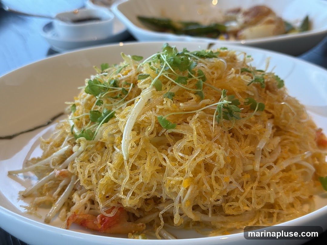 A close-up shot of a dish featuring a mound of crispy, golden noodles mixed with bean sprouts and garnished with microgreens. Hints of red seafood, possibly shrimp or crab, are visible beneath the noodles. In the blurred background, another dish and a small bowl of sauce can be seen.