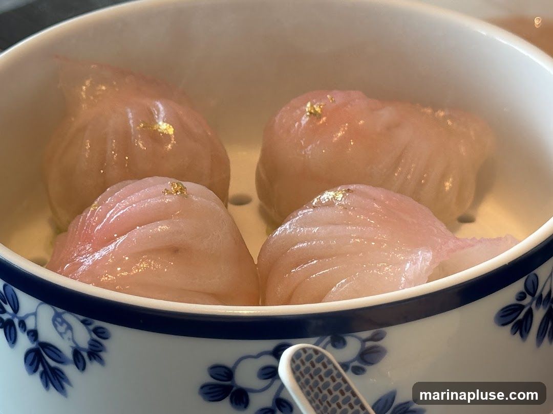 Four delicate, translucent dumplings are presented in a white ceramic steamer basket with a blue floral pattern. Each dumpling is topped with a small piece of gold leaf, adding a touch of luxury. The dumplings appear to be filled with a light-colored ingredient, possibly seafood or vegetables, and are steaming, indicating they are freshly prepared.