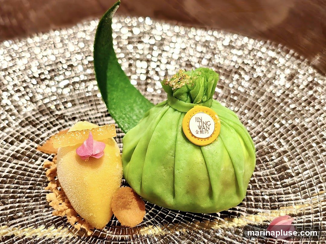 A close-up shot of a dessert presented on a textured silver plate. The dessert features a bright green, dumpling-shaped pastry with a gold leaf accent on top and a circular yellow emblem with Chinese characters and the text "JIN TING WAN". Beside the dumpling is a scoop of yellow sorbet, garnished with candied citrus peel and a small pink flower. Crumble and a small cookie are also part of the presentation. A green leaf garnish is placed behind the dumpling.