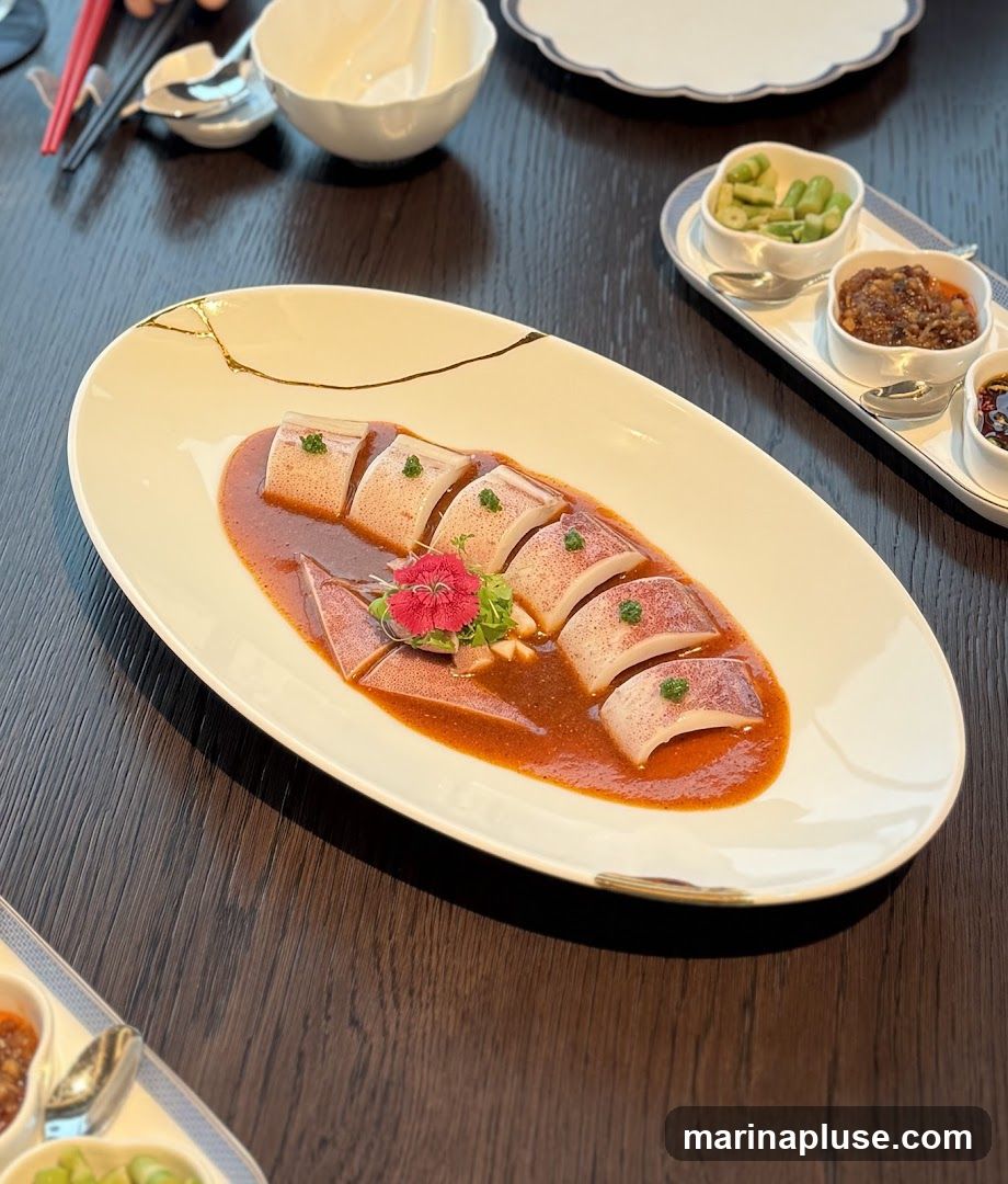 A close-up shot of a dish featuring sliced squid in a reddish-brown sauce, garnished with a small red flower and green herbs. The squid is served on an oval white plate with a gold trim. In the background, there are small bowls containing side dishes, chopsticks, and a white bowl. The table surface is dark wood.
