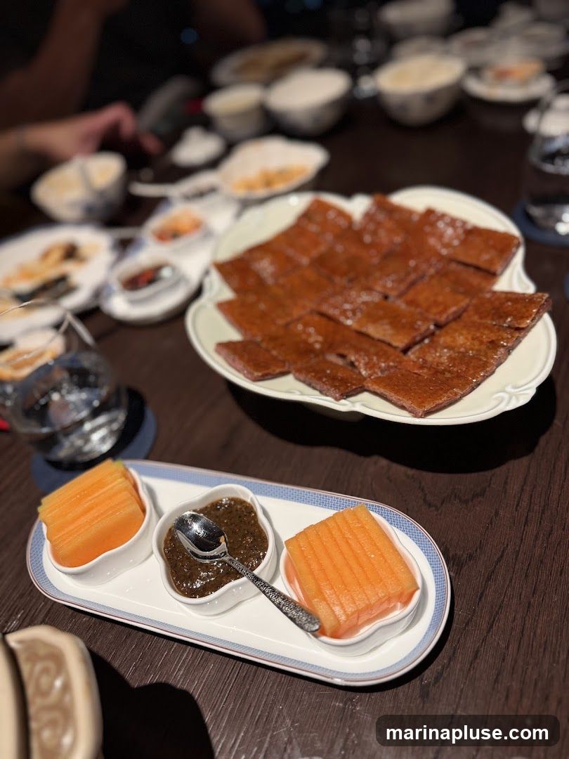 A close-up shot of a table setting with a Peking duck platter as the main focus. The duck is sliced into rectangular pieces and arranged on a white, scalloped-edge plate. In the foreground, a white tray holds two small white dishes filled with sliced cantaloupe and a dark, textured sauce, with a decorative spoon resting in the sauce. Other plates with food and drinks are visible in the blurred background, suggesting a dining experience.