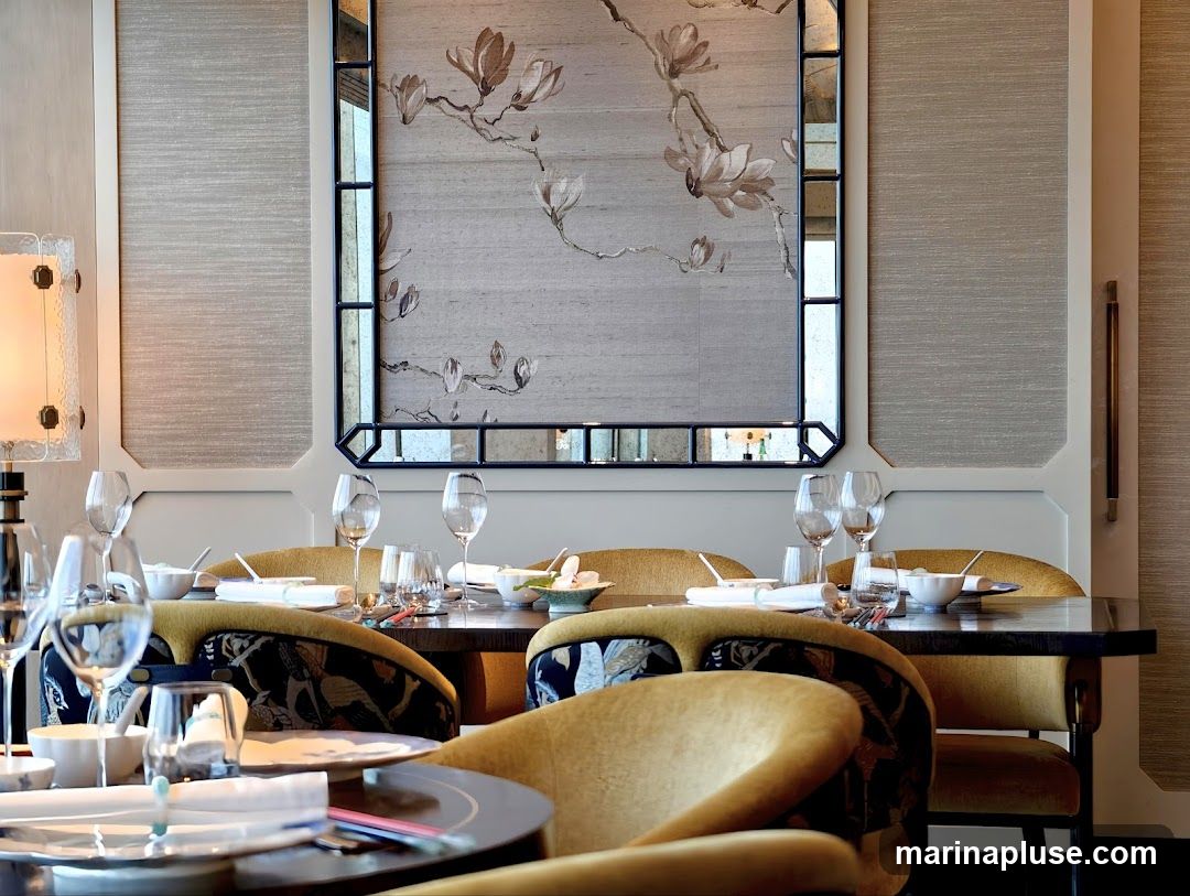 A beautifully set dining table in an elegant room. The table is set with white plates, wine glasses, and chopsticks. The chairs are upholstered in a mustard yellow fabric with a dark blue and white bird pattern. A large decorative mirror with a floral motif hangs on the wall behind the table. A lamp with a textured glass shade is visible on the left side of the image. The walls are covered in textured wallpaper.
