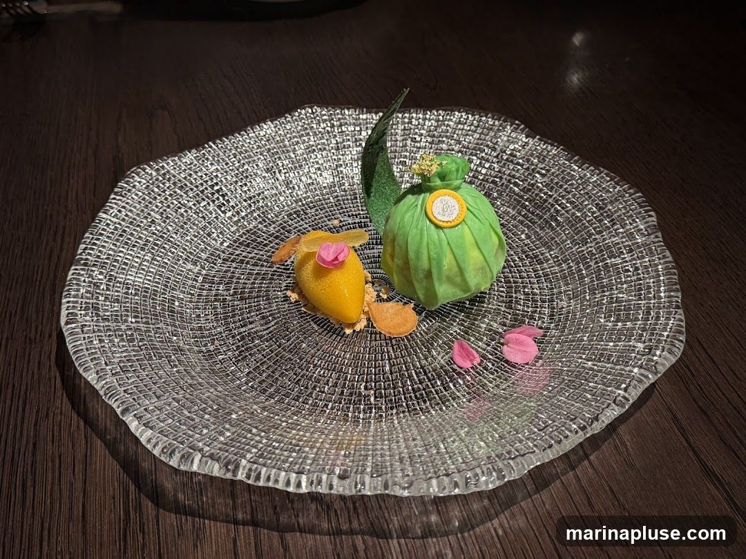 A close-up shot of a dessert served on a textured glass plate. The dessert features a bright yellow, sorbet-like scoop topped with a pink flower and a thin slice of fruit. Next to it is a green, pouch-shaped pastry adorned with gold leaf and a circular emblem. Scattered pink petals add a decorative touch. The plate rests on a dark wooden surface.