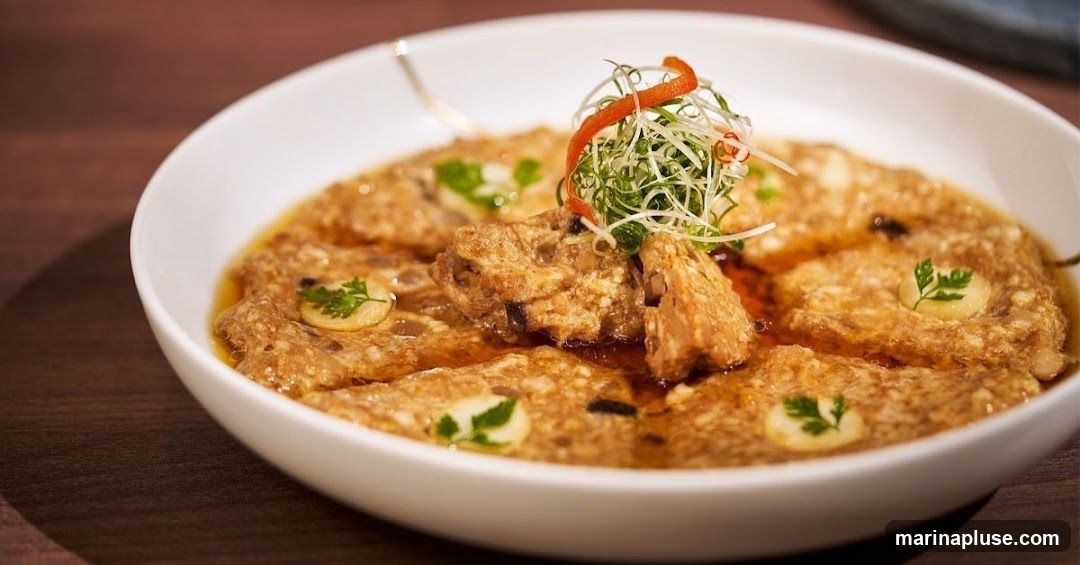 A close-up shot of a steaming dish served in a white bowl. The dish appears to be made of layered ingredients, possibly meat or tofu, covered in a savory sauce. It is garnished with finely chopped green onions, a sliver of red pepper, and small sprigs of parsley, with dollops of a creamy sauce scattered throughout. The dish is presented on a dark wooden surface.