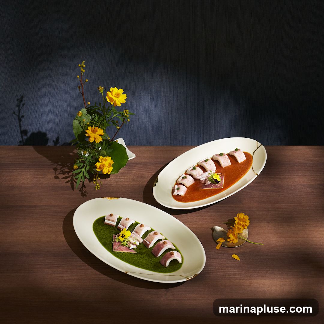 Two oval dishes are presented on a wooden table. The dish in the foreground contains sliced food arranged over a vibrant green sauce, garnished with a small piece of pink food and a yellow flower. The dish in the background holds similar sliced food over a reddish-orange sauce, with a small square garnish and a yellow flower. A vase with yellow flowers and greenery sits to the left. A small decorative bird and fallen yellow petals are to the right. The background is a dark textured wall with dappled light.
