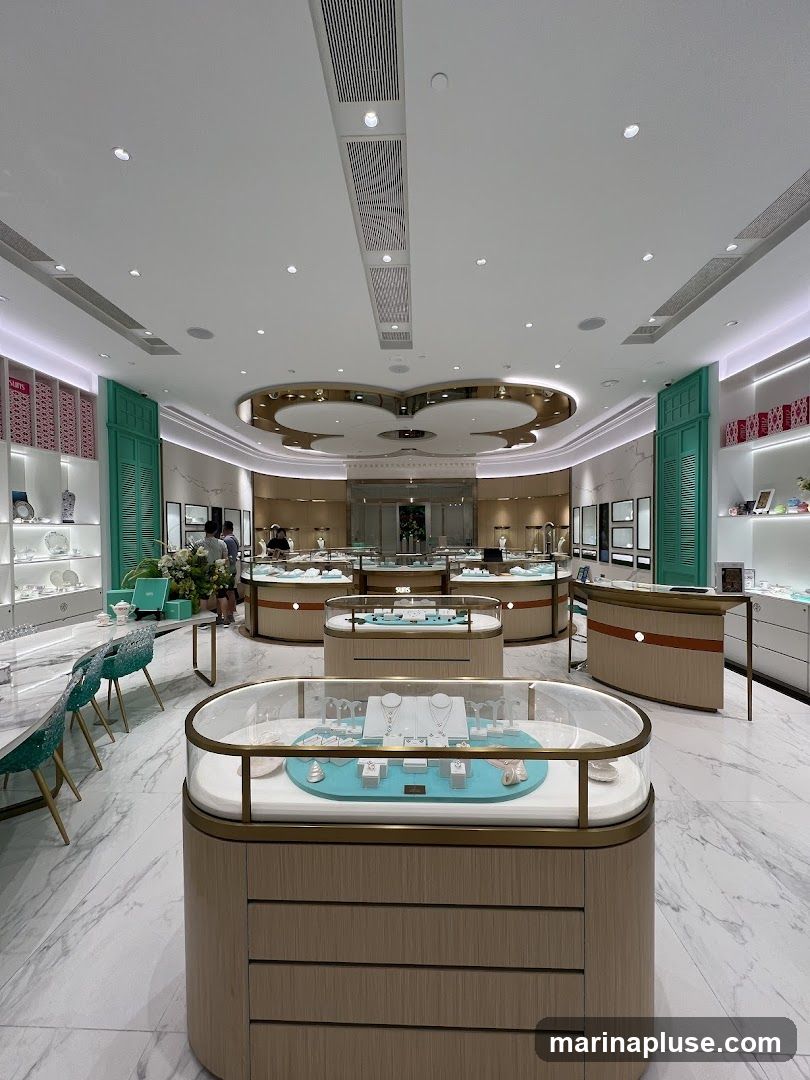 The interior of a modern jewelry store with display cases showcasing necklaces, bracelets, and earrings. The store features a clean, elegant design with marble flooring, light wood accents, and teal seating. Several customers are browsing the merchandise.