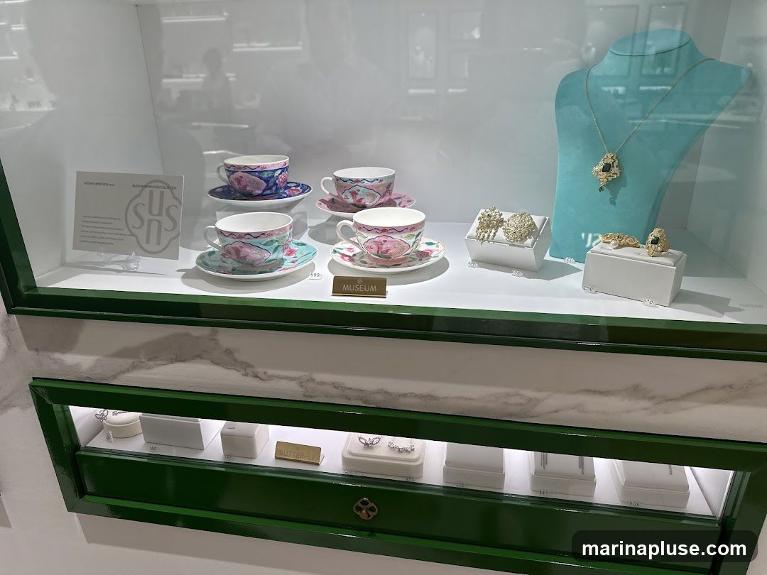A display case featuring decorative teacups and saucers, along with various pieces of jewelry including a necklace, earrings, a ring, and a bracelet. The teacups are adorned with floral and bird motifs. The jewelry includes intricate designs, some with gemstones. A label below the teacups reads "MUSEUM". Another label on the left side has text and a logo. The lower section of the display case shows more jewelry items in individual boxes.