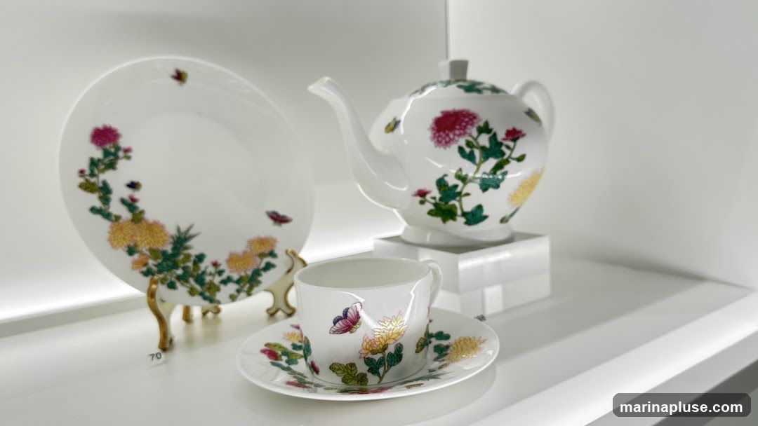 A white porcelain teapot, cup, saucer, and plate are displayed on a white shelf. The set is adorned with colorful floral and butterfly motifs. The plate is propped up on a stand.