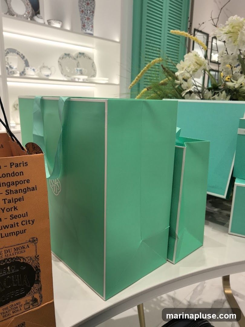 Several teal-colored shopping bags with white trim are displayed on a white surface. One bag on the left is partially obscured by an orange paper bag with text that includes 'MOKA COFFEE'. In the background, shelves display porcelain items, and a floral arrangement is visible.
