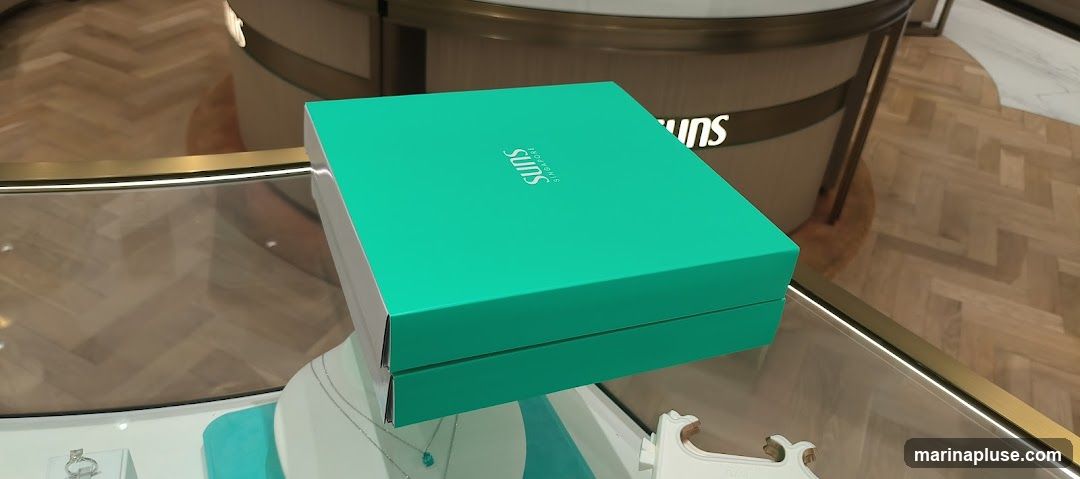 A close-up shot of a teal-colored jewelry box with the brand name 'Suns Singapore' printed in white on its lid. The box is placed on a glass display counter, reflecting the herringbone-patterned wooden floor and the interior of a store. Below the box, a white mannequin bust displays a delicate silver necklace with a blue pendant. Another display case to the left shows a ring. In the background, a curved counter with the 'Suns' logo is visible.