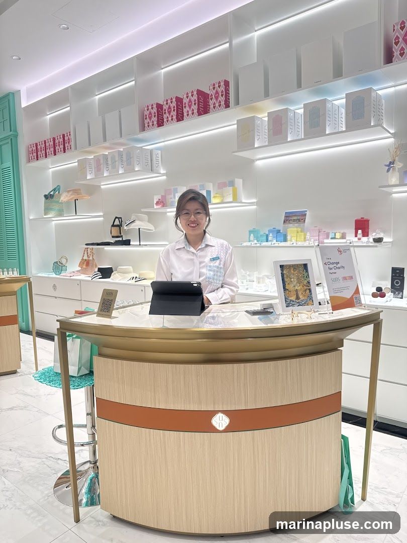 A woman in a white collared shirt stands behind a wooden counter in a brightly lit store. The shelves behind her are stocked with various products, including boxes, hats, and small decorative items. A tablet is on the counter in front of her.