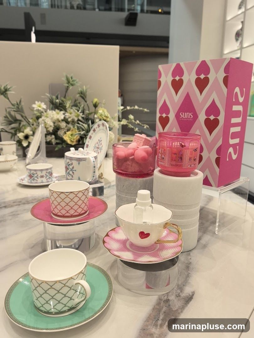 A display of decorative teacups and saucers, some with intricate patterns and gold accents. In the background, there are floral arrangements and pink decorative boxes with heart patterns. A small bottle of hand sanitizer is placed in one of the teacups.