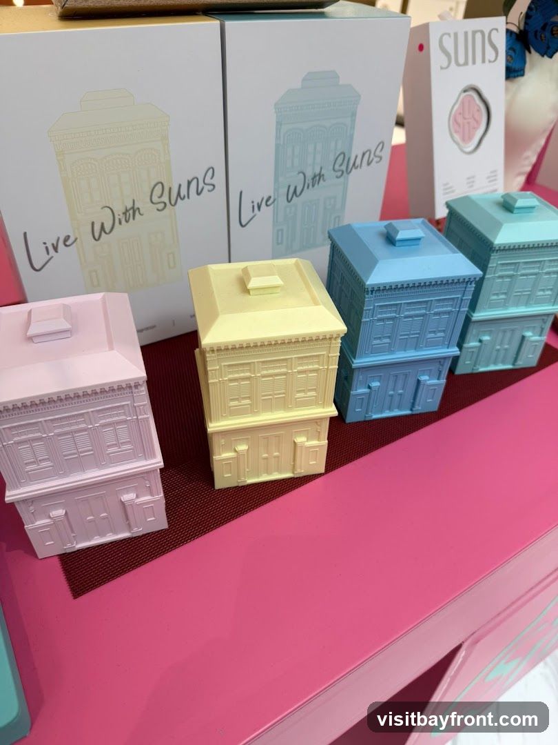 A display of miniature house-shaped objects in pastel colors: pink, yellow, light blue, and teal. These are arranged on a pink surface, with boxes in the background featuring the text "Live With Suns" and an illustration of a similar house. A product box with the brand name "Suns" is also visible.