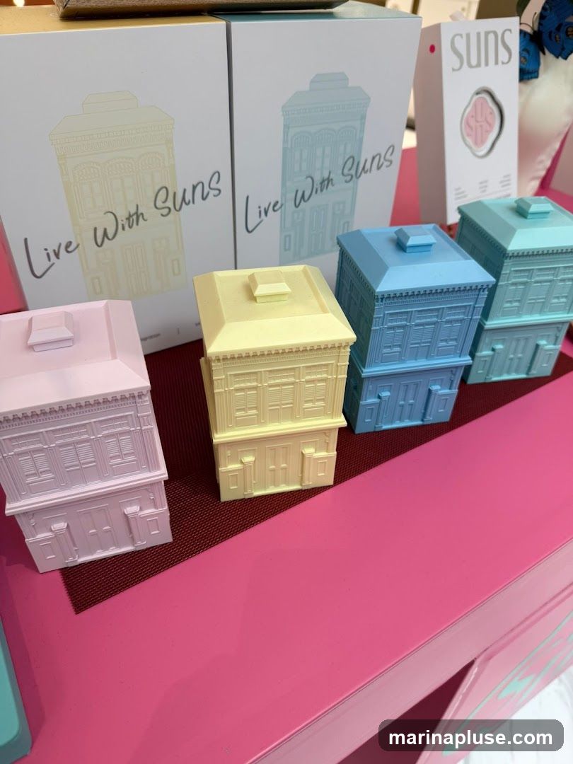 A display of miniature house-shaped objects in pastel colors: pink, yellow, light blue, and teal. These are arranged on a pink surface, with boxes in the background featuring the text "Live With Suns" and an illustration of a similar house. A product box with the brand name "Suns" is also visible.