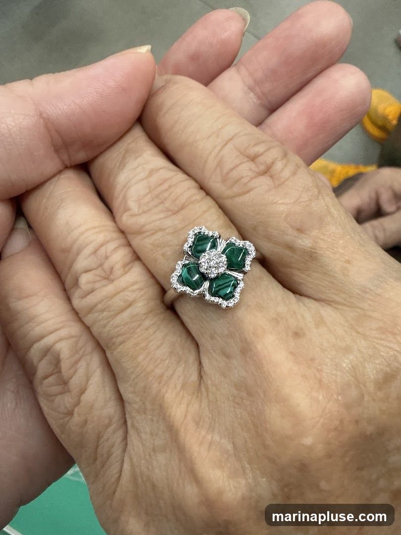 A close-up shot of a ring on a person's finger. The ring is designed as a flower with malachite petals and a center of small diamonds. The malachite has a distinctive banded green pattern. The ring is set in white metal, likely silver or white gold, and is further embellished with small, sparkling diamonds around the edges of the petals. The background is softly blurred, with hints of skin tones and possibly fabric.