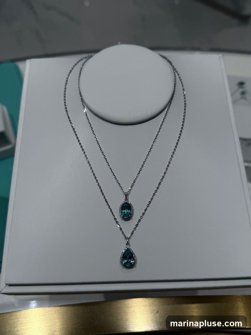 Two silver necklaces are displayed on a white stand. The top necklace has a small oval-shaped blue pendant surrounded by small clear stones. The bottom necklace has a larger pear-shaped blue pendant, also surrounded by small clear stones. The background is blurred, but appears to be a store interior with a turquoise accent.