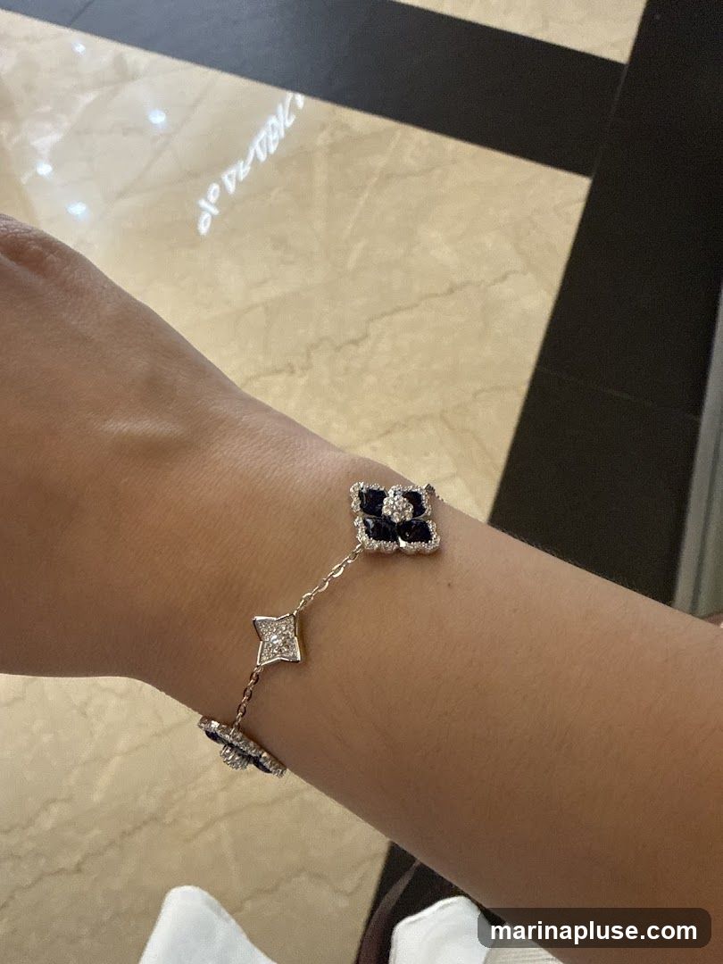 A close-up shot of a person's arm wearing a delicate bracelet. The bracelet features a chain with several charms. The charms are shaped like four-leaf clovers or flowers, adorned with small diamonds and dark blue stones, likely sapphires. The background shows a marble floor with dark stripes.